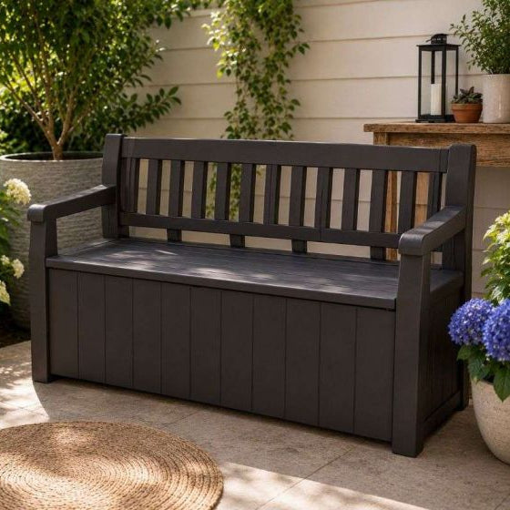 Dark charcoal gray outdoor storage bench with a slatted backrest, set in a garden patio environment with plants, a woven rug, and a wooden table in the background.